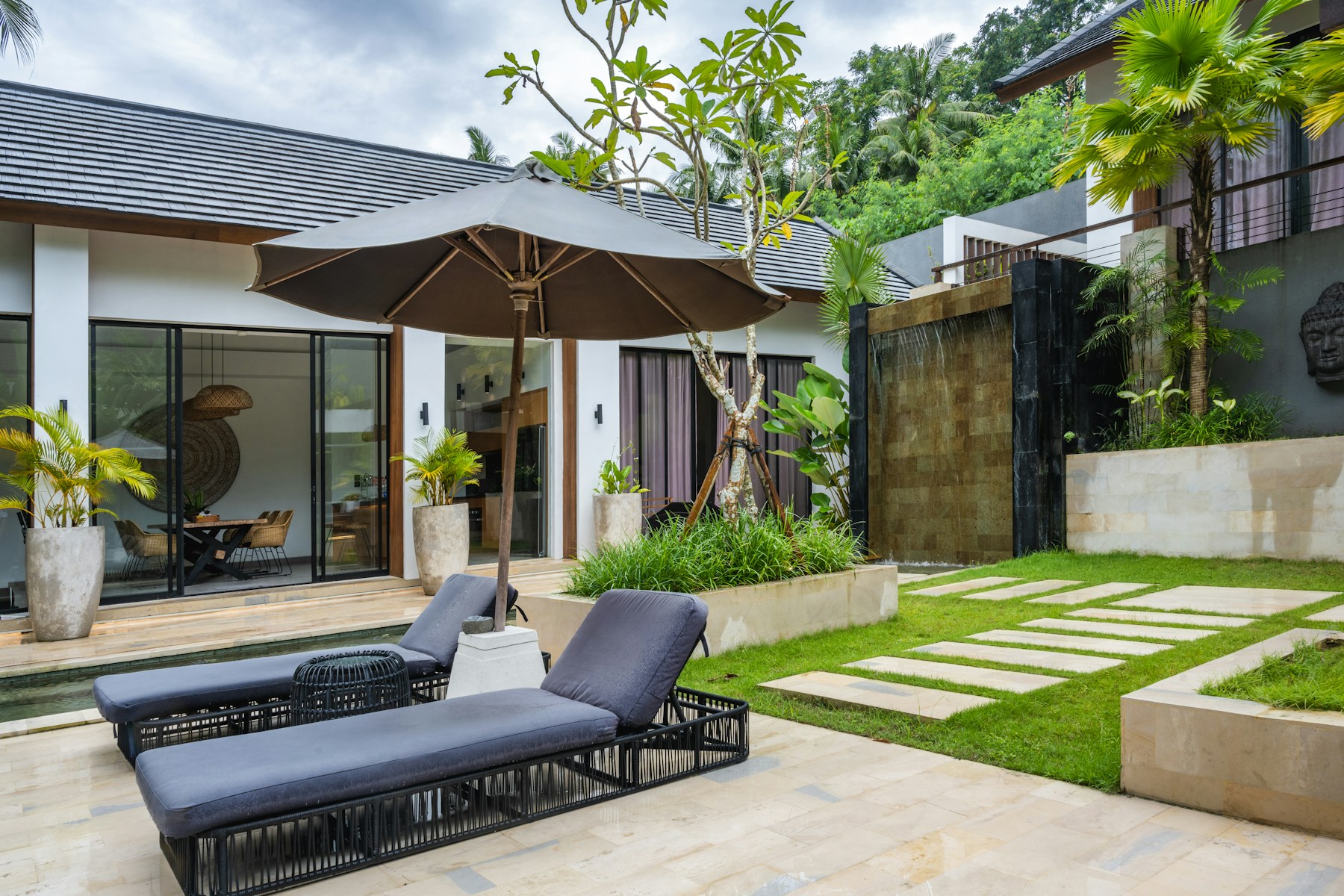 A lush courtyard with a stone path, water wall, and shaded lounge
